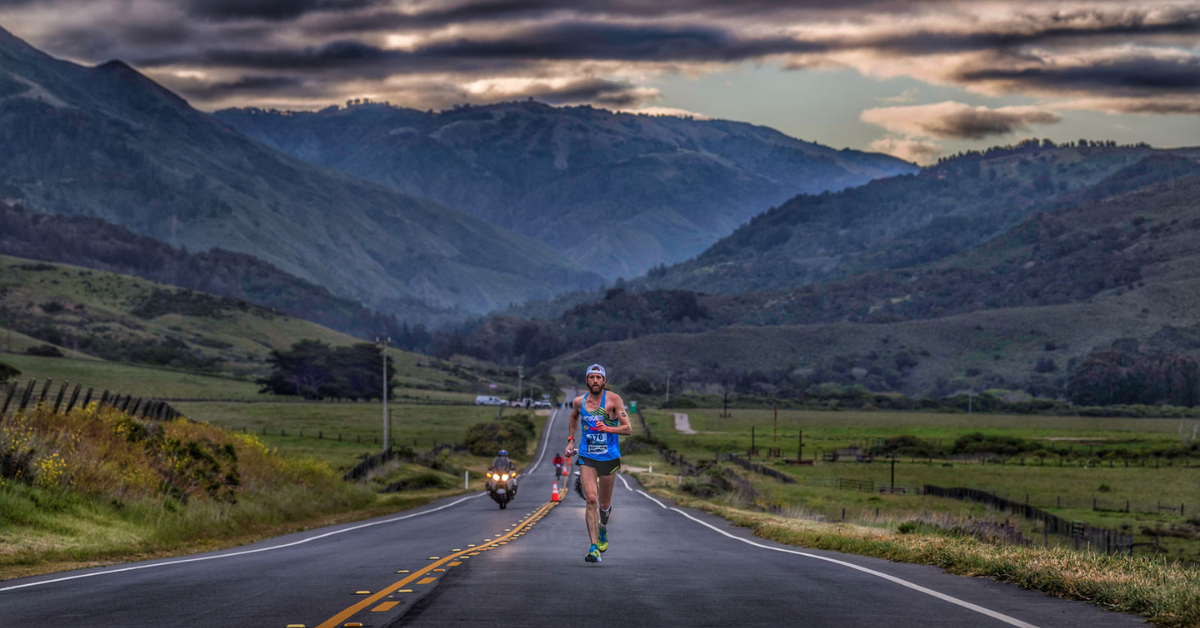 Runner with mountains in backgrouns faces camera as he runs along highway