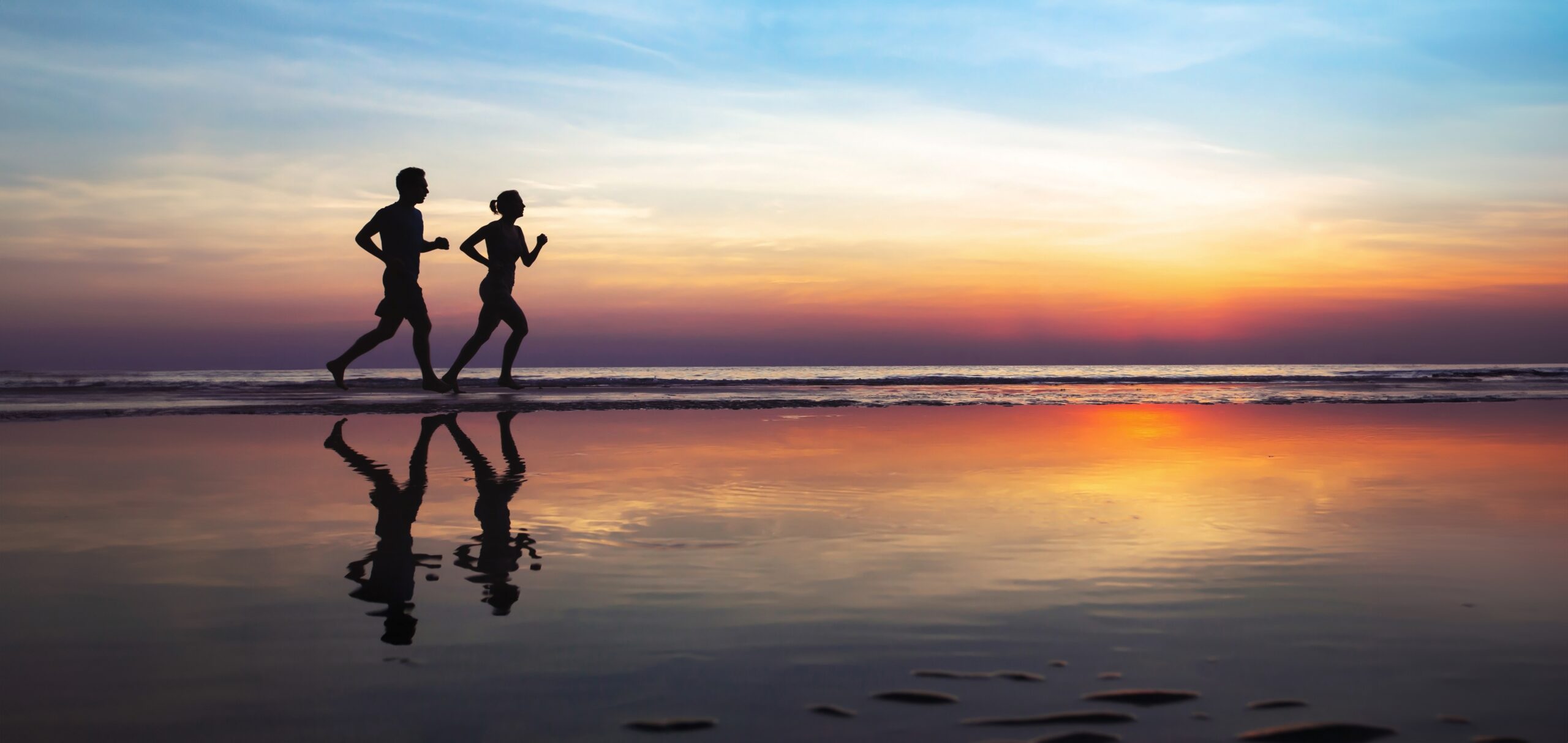 Silhouettes of two runners, a woman and a man, running on a beach by the ocean after sunset, enjoying the health benefits of running