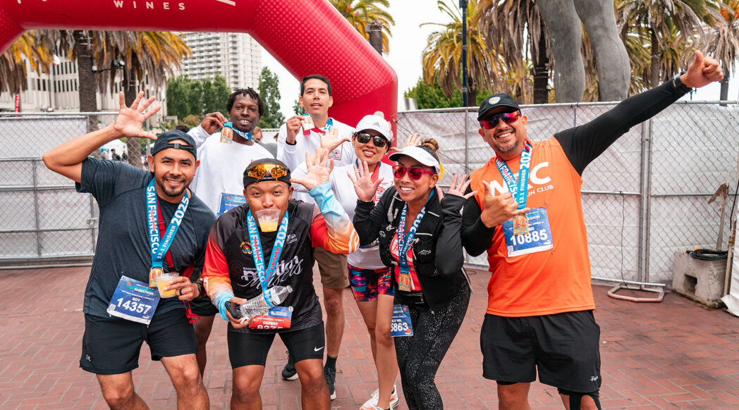 A joyful group of seven marathon finishers pose together under a red FitVine Wines arch at the San Francisco Marathon Finish Line Festival.