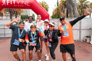 A joyful group of seven marathon finishers pose together under a red FitVine Wines arch at the San Francisco Marathon Finish Line Festival.