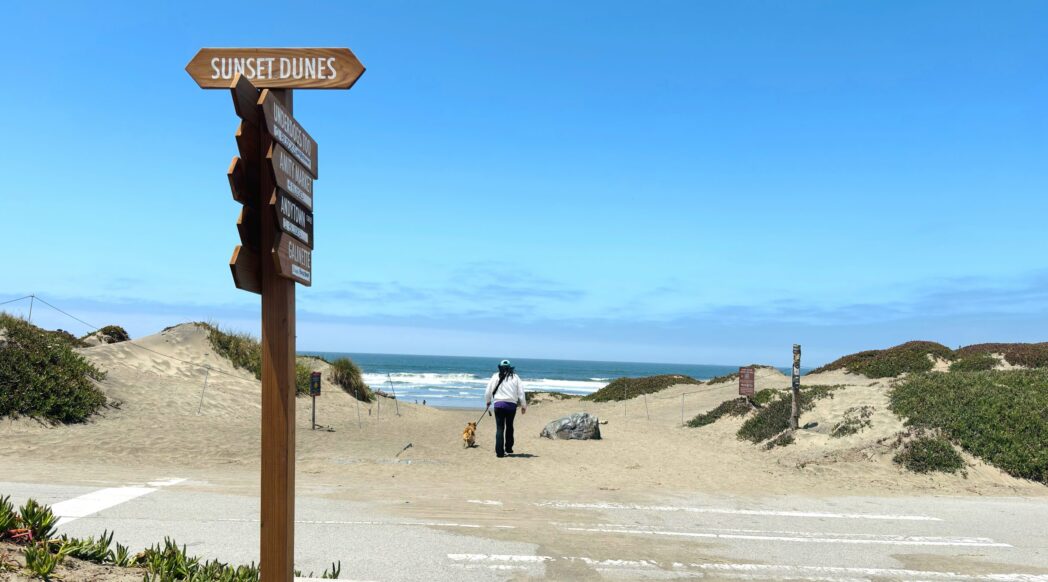 A view of Sunset Dunes with the Pacific ocean in the background and a sign pointing toward the park in the foreground