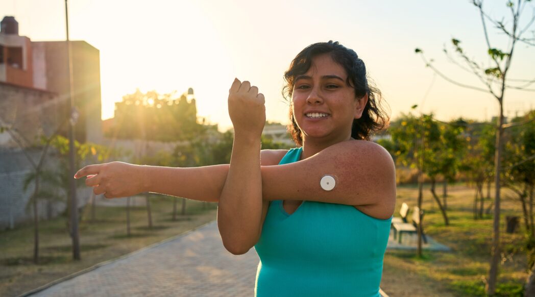 A Latina runner with a Continuous Glucose Monitor stretches before her run with sunrise in the background