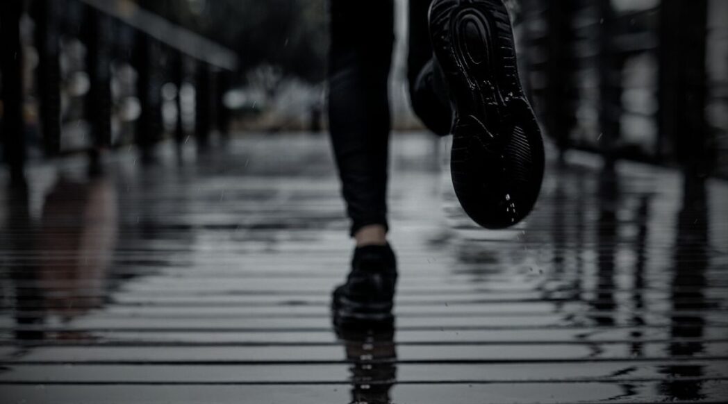 Close-up of a person running on a wet wooden boardwalk in the rain, with water reflections and motion blur on their black sneakers and legs.