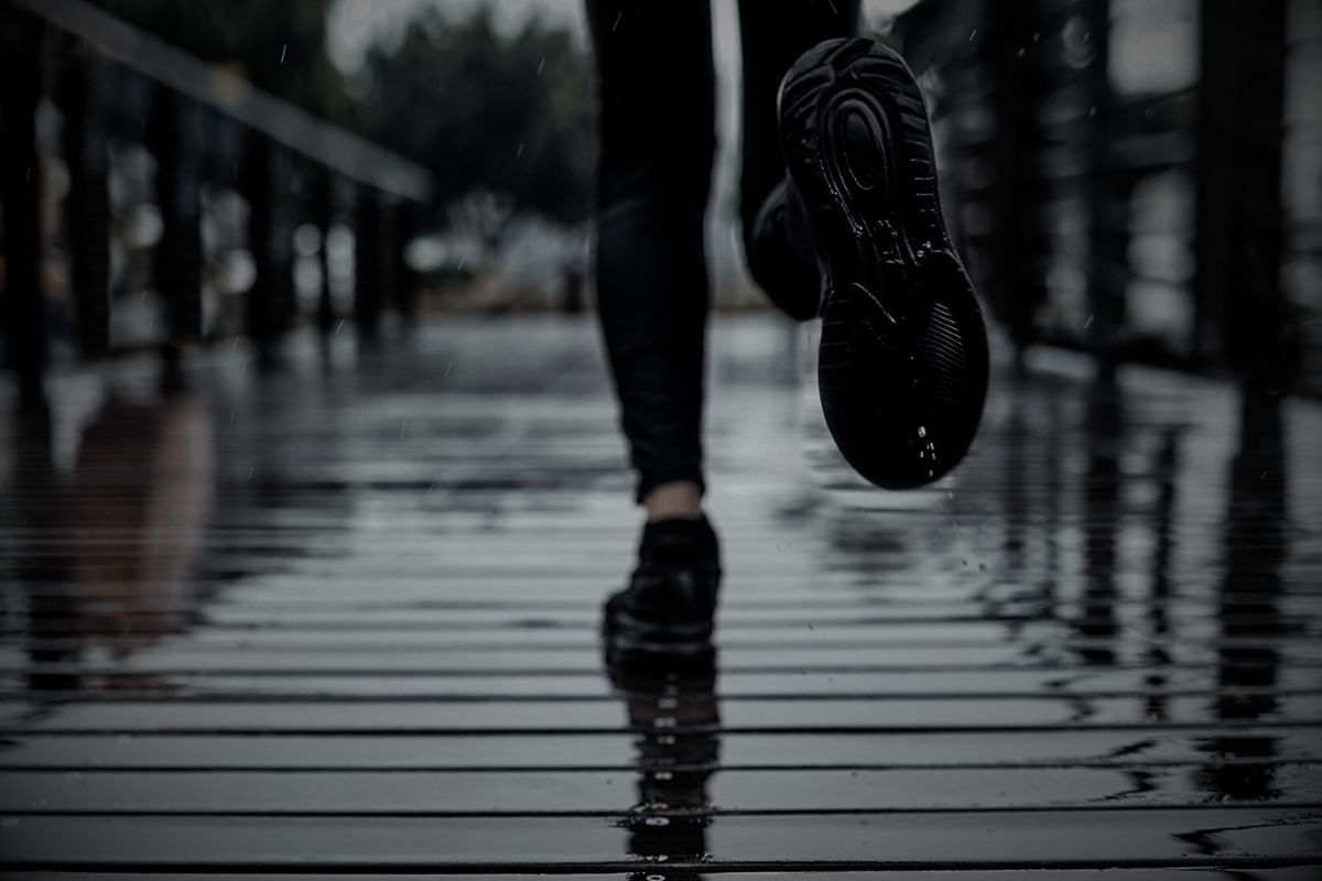 Close-up of a person running on a wet wooden boardwalk in the rain, with water reflections and motion blur on their black sneakers and legs.