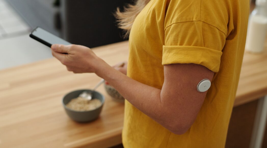 A close-up on a woman with a yellow shirt wearing a continuous glucose monitor and tracking her meal