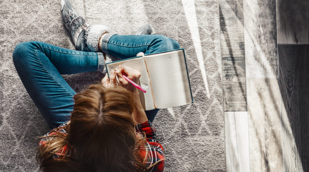 A woman is sitting cross-legged on the floor wearing woolen socks and a chunky sweater and journaling, doing her 2025 reflection