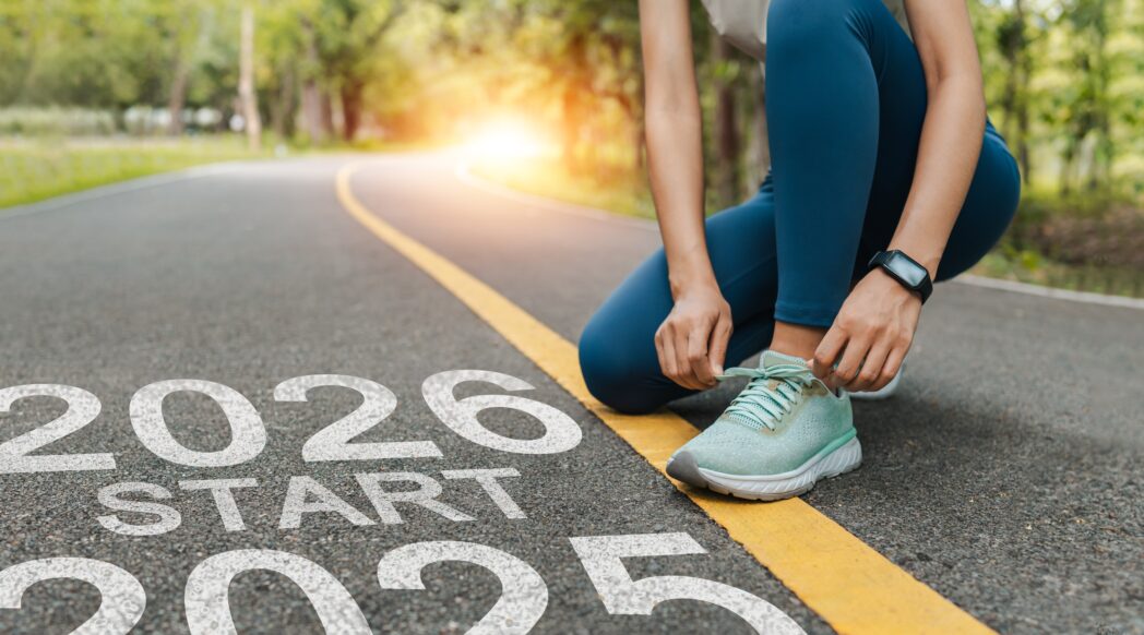 A woman is tying her running shoes on a road with a 2026 sign, ready to achieve her running goals