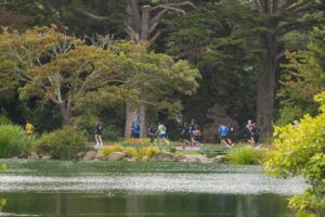A group of people run on a road behind a scenic lake with huge trees in the background in Golden Gate Park