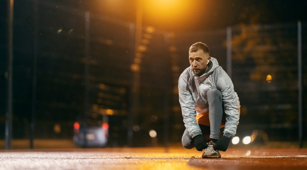 A man is getting to run in the dark on a well-lit empty street, tying his shoes.